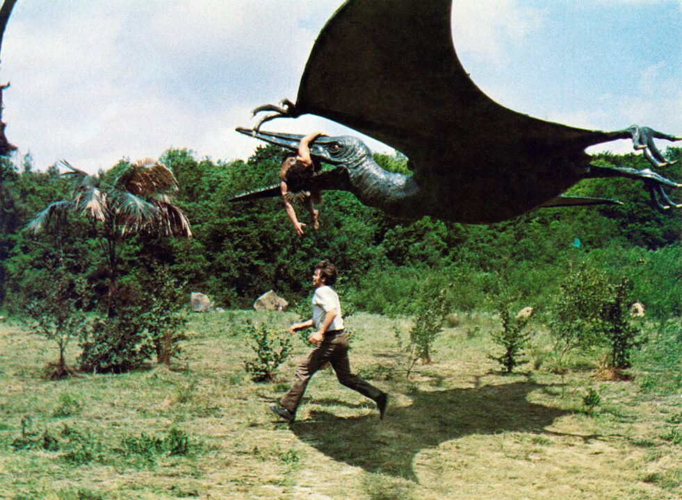 A scene from the 1974 movie "The Land That Time Forgot." It depicts a flying dinosaur, Pteranodon, swooping low to the ground and picking up a caveman in its jaws and beginning to fly away, as a modern man, playing by Doug McClure, runs just below it in an effort to stop it.