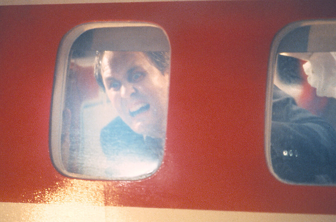 a scene from the fourth segment of "Twilight Zone: The Movie," an adaptation of "Nightmare at 20,000 Feet." It is a closeup looking from the outside of an airplane in the window. Looking out of that window is John Lithgow as a passenger with his face pressed up against the glass with a terrified expression on his face, and looking to be screaming at whatever he is seeing out on the plane's wing.