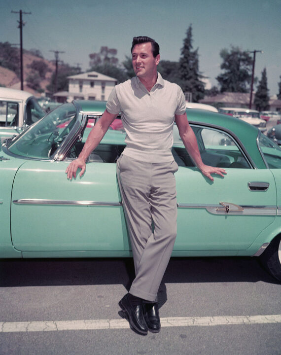American actor Rock Hudon (1925 - 1985) leans against a sea foam green car in a parking lot, 1950s.