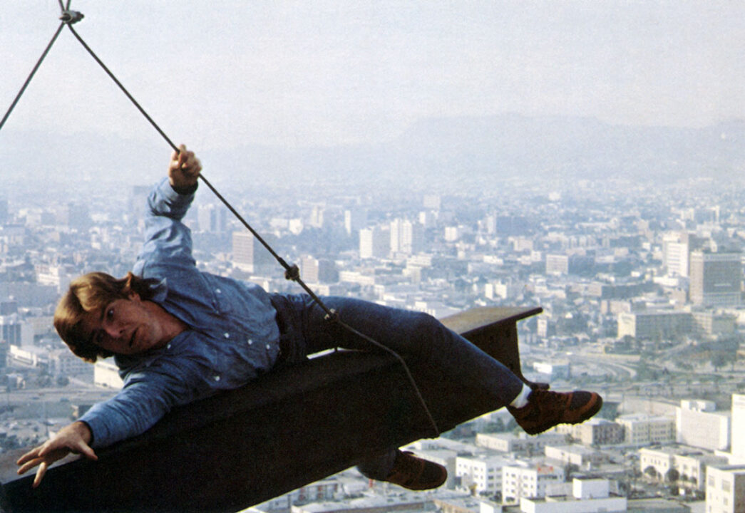 scene from the 1984 movie "Dreamscape." It depicts Dennis Quaid's character psychically projected into a patient's nightmare. He is high above a city on a suspended construction girder. He is looking scared as he lays on the girder, seeming as if he is trying to keep himself from falling off.