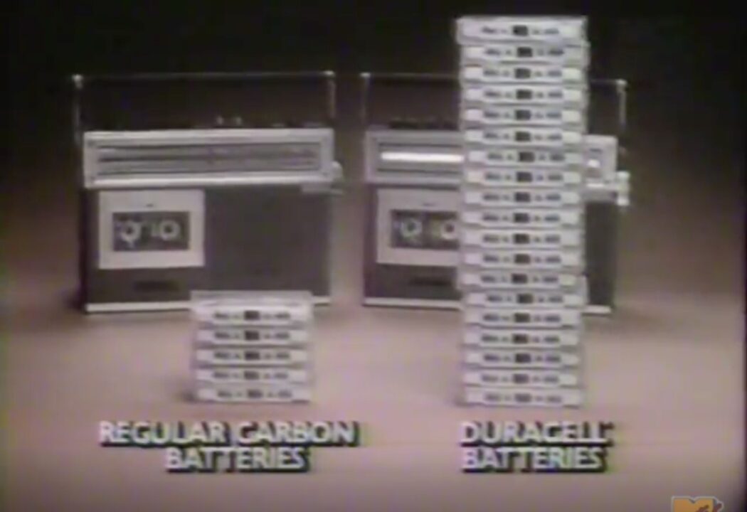screenshot of a 1983 Duracell battery commercial. Pictured in the background are two boomboxes. In front of the one on the left is a small pile of cassette tapes, over which is yellow text reading "Regular Carbon Batteries." In front of the boom box on the right is a much larger stack of tapes, with lettering in front of it reading "Duracell Batteries."