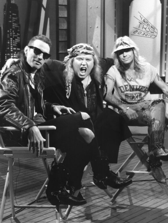 black-and-white still from a 1990 episode of NBC's "Friday Night Videos." Seated from left to right are that episode's guest hosts, Tommy Lee, Sam Kinison and Vince Neil