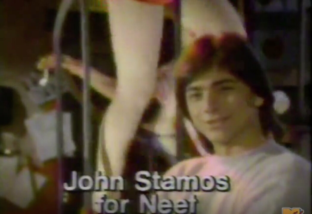 screenshot from a 1983 commercial for a Neet hair removal product. A young John Stamos is pictured in medium closeup, in front of a woman whose legs can only be seen. Yellow text reads: "John Stamos for Neet"