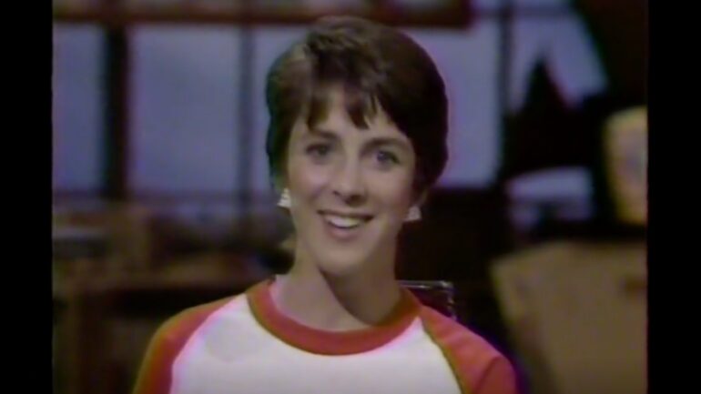 screenshot from a July 16, 1983, broadcast on MTV. The shot is a medium closeup of VJ Martha Quinn, who is wearing a shirt colored white below the neck, with red ring around the neck and red shoulders down into the arms.