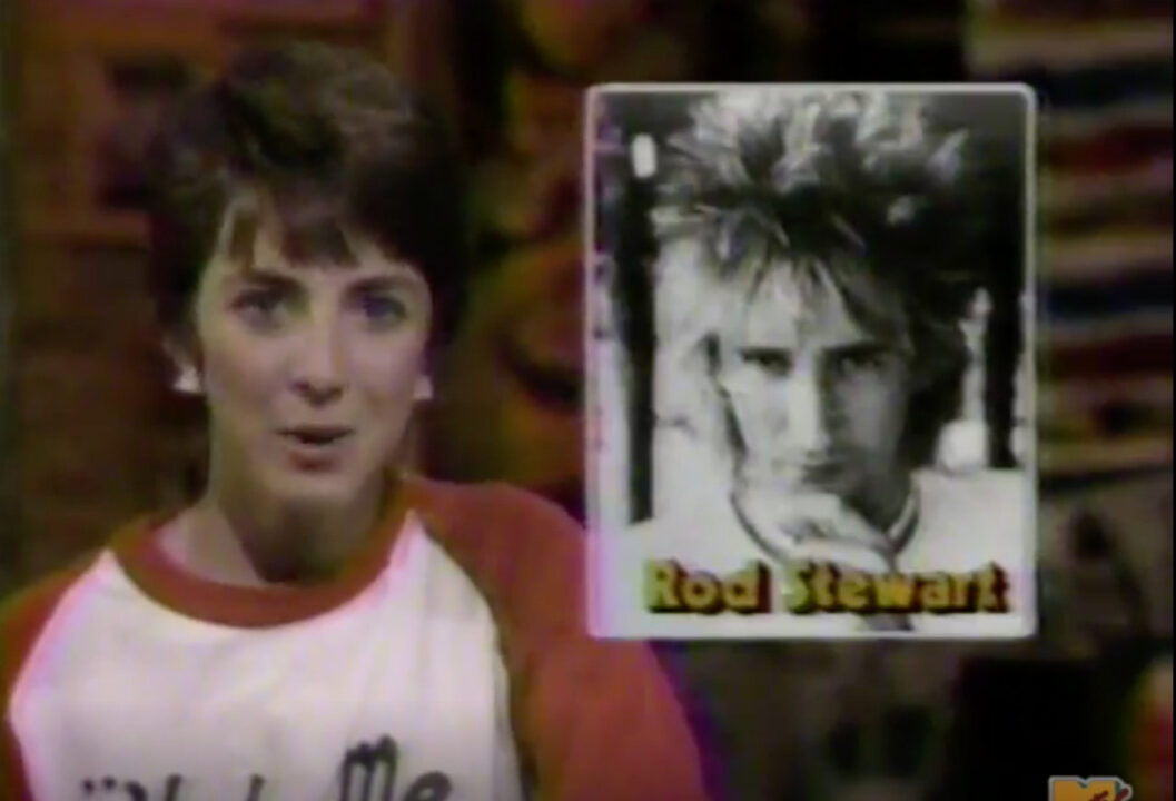 screenshot from a July 16, 1983, MTV broadcast. It is a medium closeup of VJ Martha Quinn as she reports on news about Rod Stewart, who is pictured in a large black-and-white image to the left of Quinn, who is seated in a chair.
