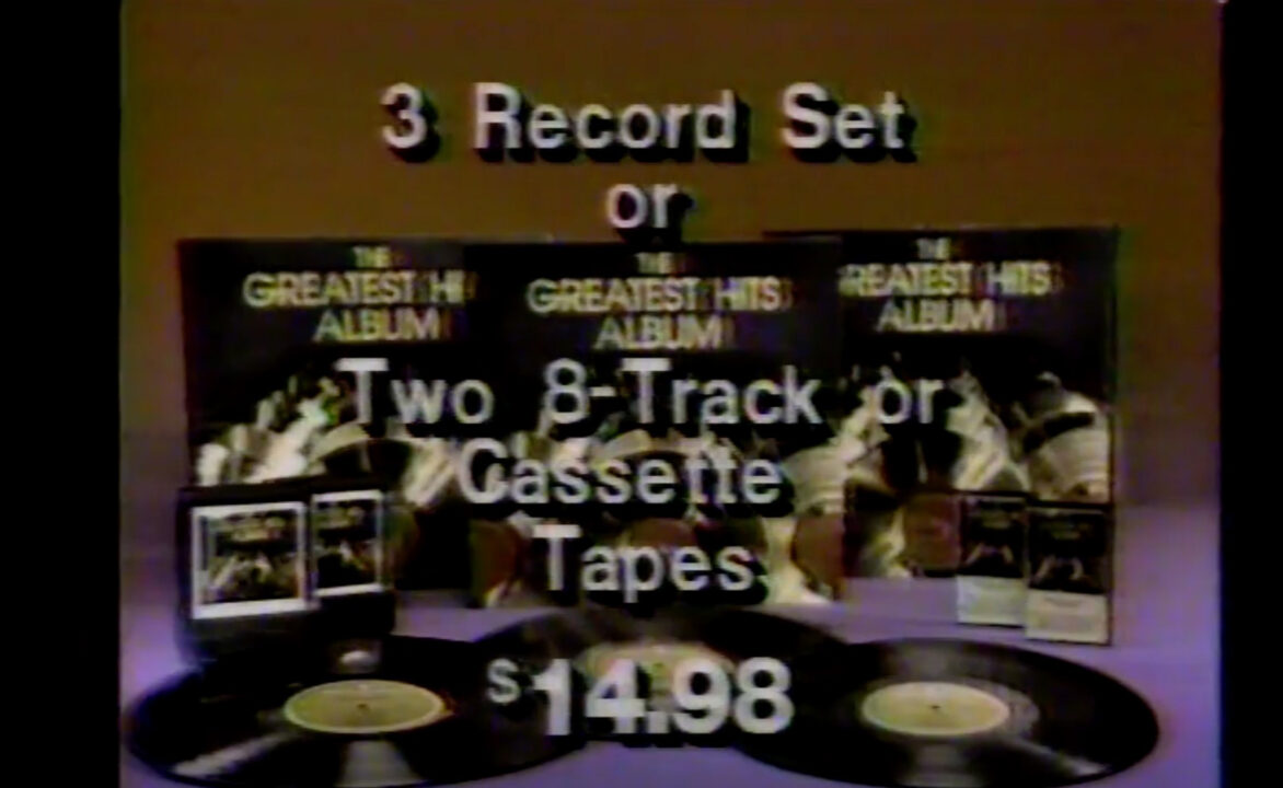 screenshot from a 1983 commercial for "The Greatest Hits Album" collection from Sessions Records. The image has the vinyl record album covers stacked across the background, with the actual records laying in front of each jacket. There are also cassette tapes of the collection, and 8-tracks. White text reading down over the imagery says: "3 Record Set or Two 8-Track or Cassette Tapes $14.98"