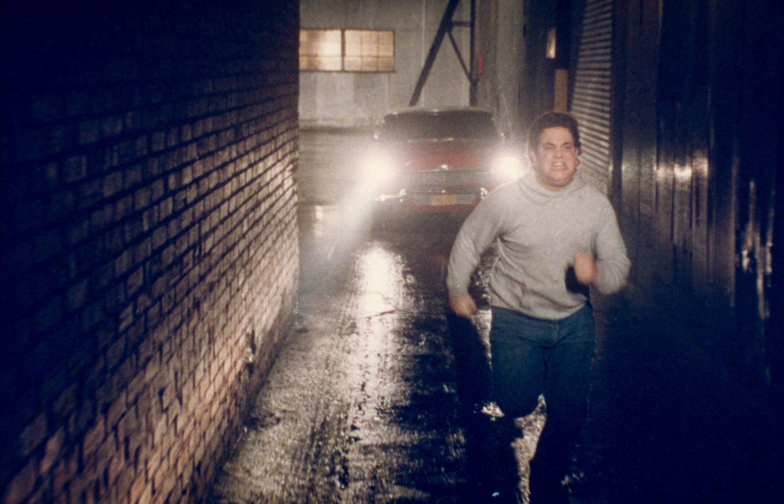 photo from the 1983 movie "Christine." It pictures Malcolm Danare as "Moochie," one of the characters who had been bullying Arnie, who owns the haunted 1958 Plymouth Fury named Christine. Christine is now, in this scene, chasing Moochie down a very narrow, dark alley, headlights beaming as the heavier-set Moochie desperately runs with terror in his eyes.