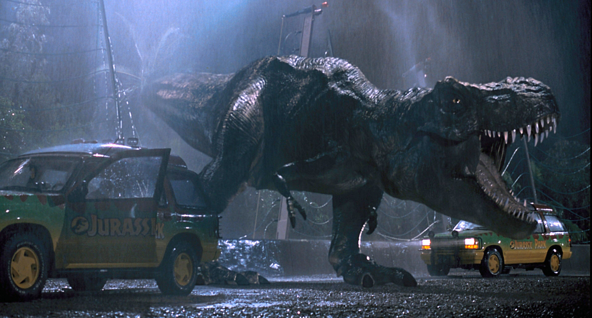 photo from the 1993 movie "Jurassic Park." It depicts the T. rex attacking two of the Jurassic Park tour trucks as it breaks through a fence, roaring, at night during a rainstorm.