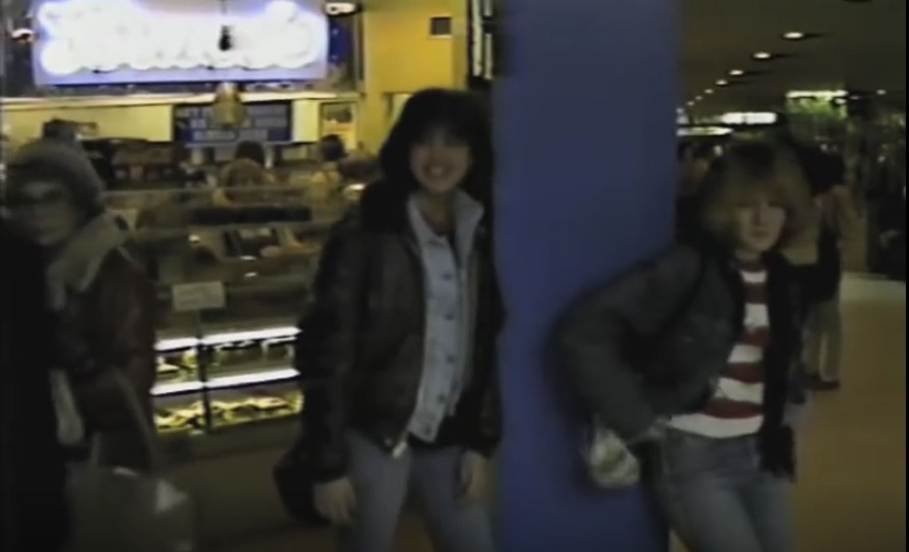 screenshot from the 1983 video documentary "Mall City." It pictures two teenage girls hanging out at a Long Island shopping mall, laughing as they stand in front of a cookie/pastry store