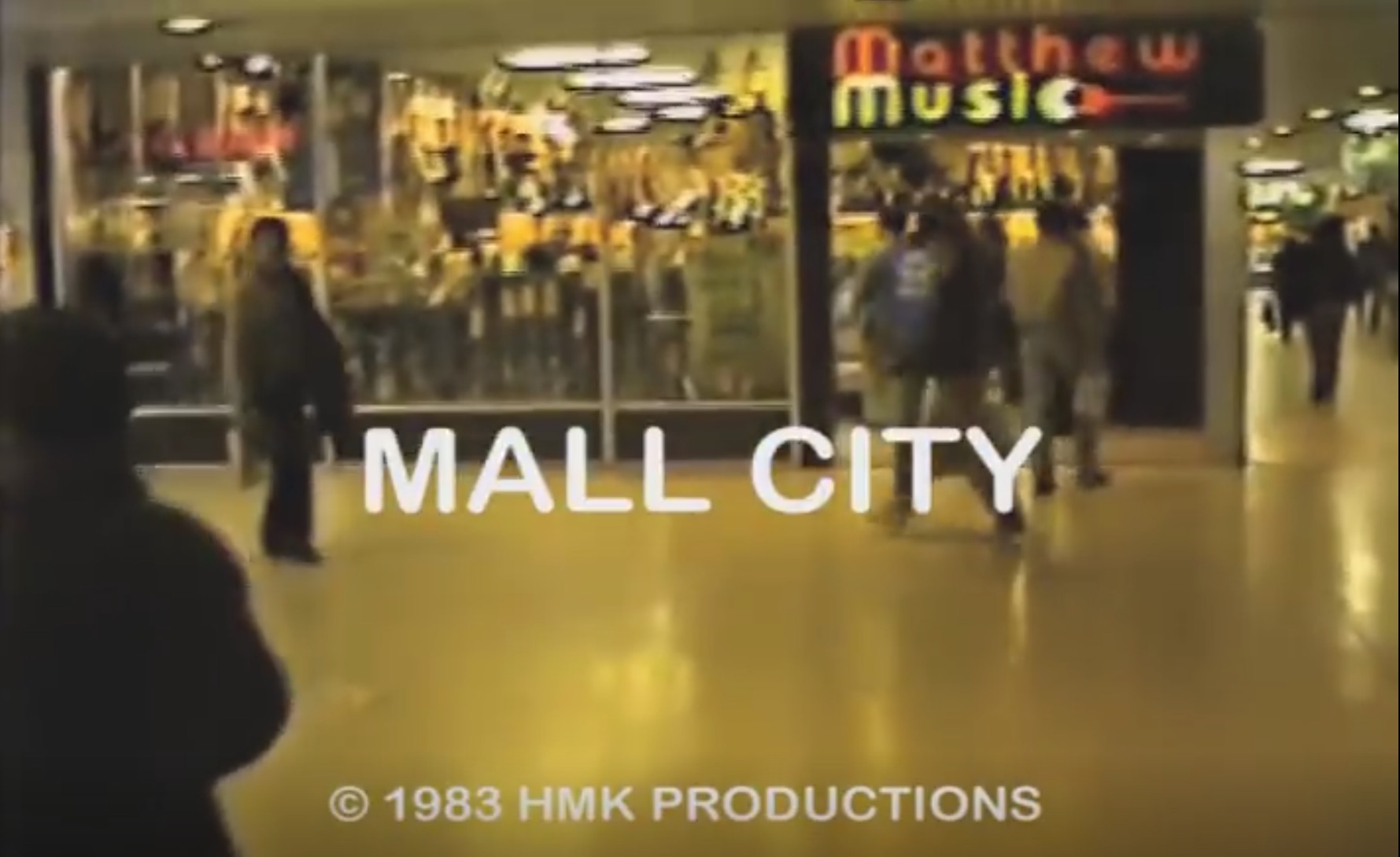 screenshot of the title credit screen from the 1983 video documentary "Mall City." It is a shot inside a Long Island shopping mall, with people walking in front of a store called Matthew Music. The title, "Mall City," reads in white lettering over the scene. Below that is the text "© 1983 HMK Productions"
