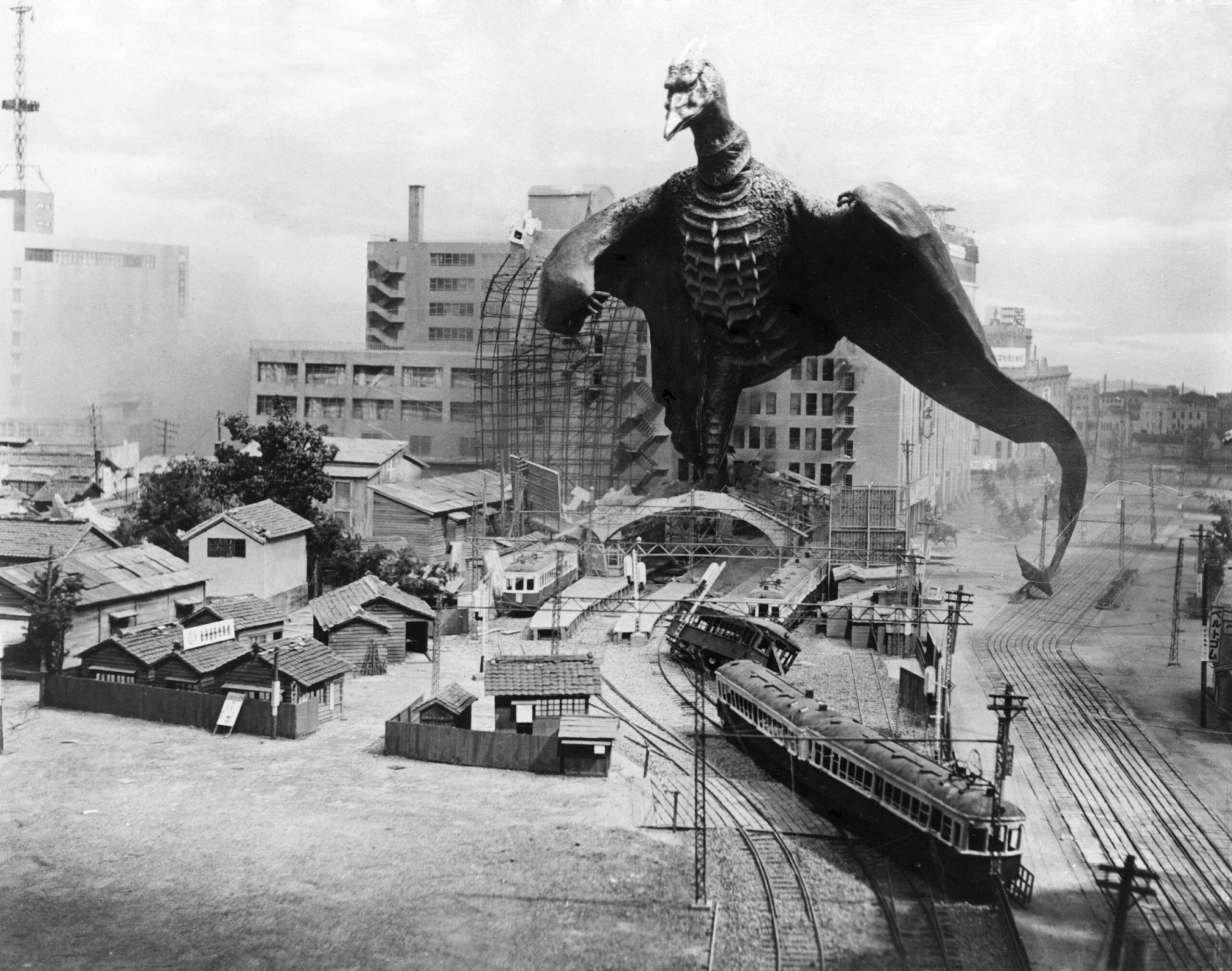 black and white photo from the 1956 Toho film "Rodan." It depicts the title monster, a giant, Pterandon-like flying creature, having landed in a city, where it is demolishing buildings as it walks along.