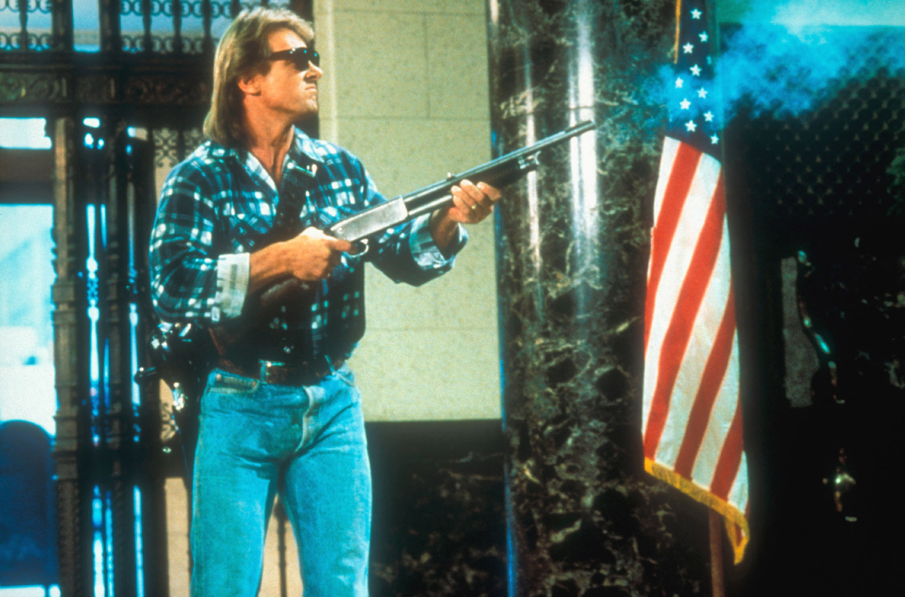 scene from the 1988 movie "They Live." Roddy Piper's character, Nada, has entered a building and is standing next to an American flag. He is wearing a plaid shirt, jeans and dark sunglasses and firing a rifle at the aliens he is able to see with those glasses.