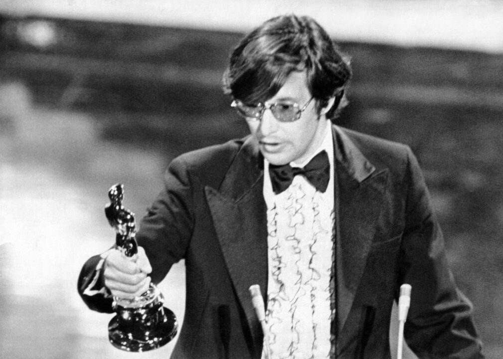 black and white photo of William Friedkin accepting his Best Director Oscar for "The French Connection" at the 1972 Academy Awards ceremony. He is wearing glasses and a 70's-era tuxedo, with white frilled shirt, and is holding the Oscar statuette out in his right hand as he gives a speech at the podium.