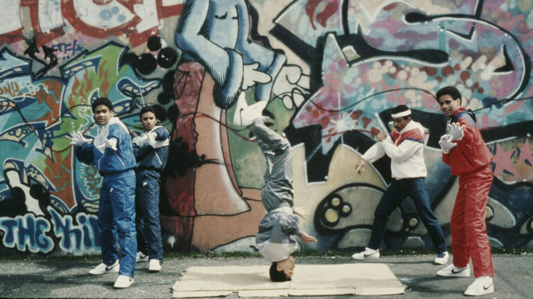 Teenagers breakdancing next to a wall covered in grafitti, Brooklyn, New York, April 1984.