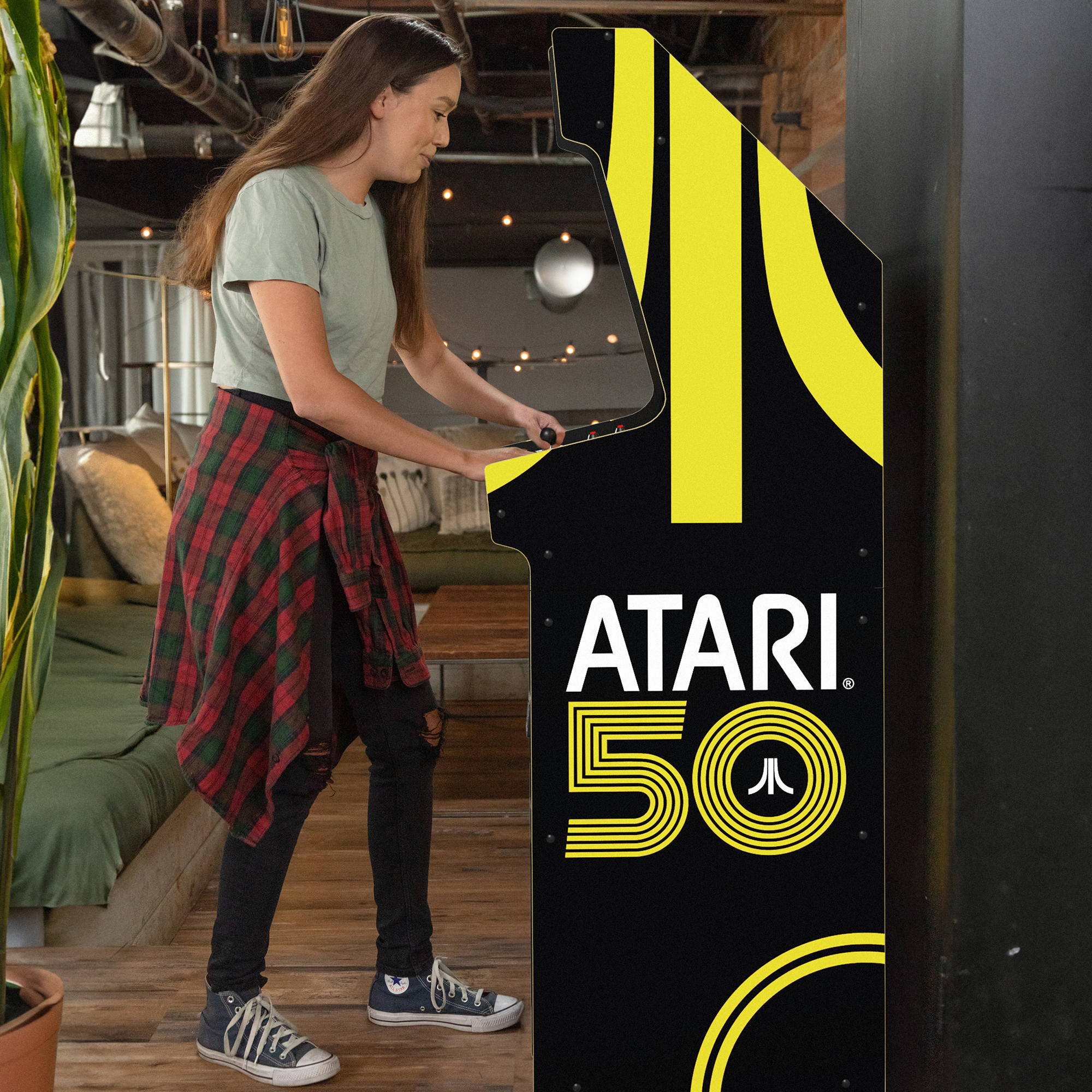 image of a young woman playing the Atari 50th Anniversary Deluxe Machine from Arcade1Up. She and the arcade cabinet are seen from the side; she is wearing grey Converse sneakers and a grey short-sleeved shirt, with a flannel longer-sleeved shirt wrapped around her waist as she plays, and has a slight smile as she focuses on the screen. The side of the cabinet has part of the Atari "Fuji" logo in yellow, and lettering reads "Atari 50."