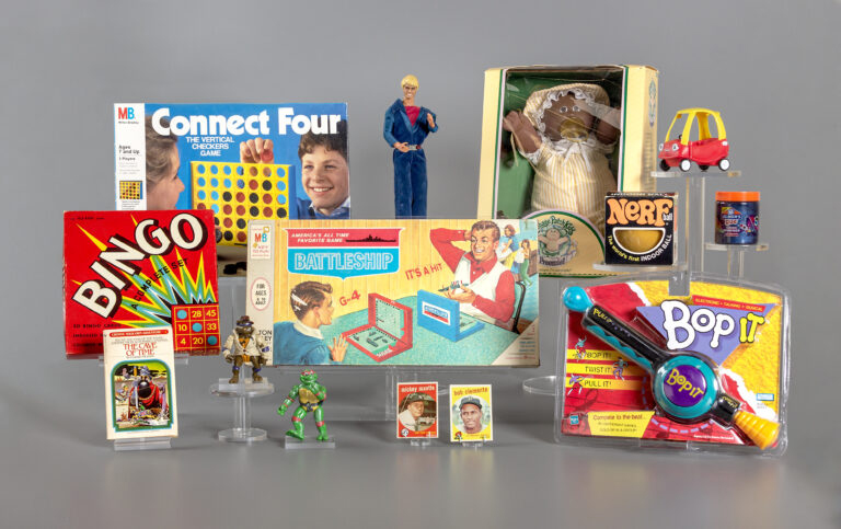 picture of the 12 finalists for 2023 induction into the National Toy Hall of Fame. Pictured are the board game Connect Four; the Ken doll; a Cabbage Patch Kid; a Little Tykes Cozy Coupe; a Nerf ball; slime; Bop It; Battleship game; a box containing a bingo game; a Choose Your Own Adventure book; decks of baseball cards; and two Teenage Mutant Ninja Turtle figures.