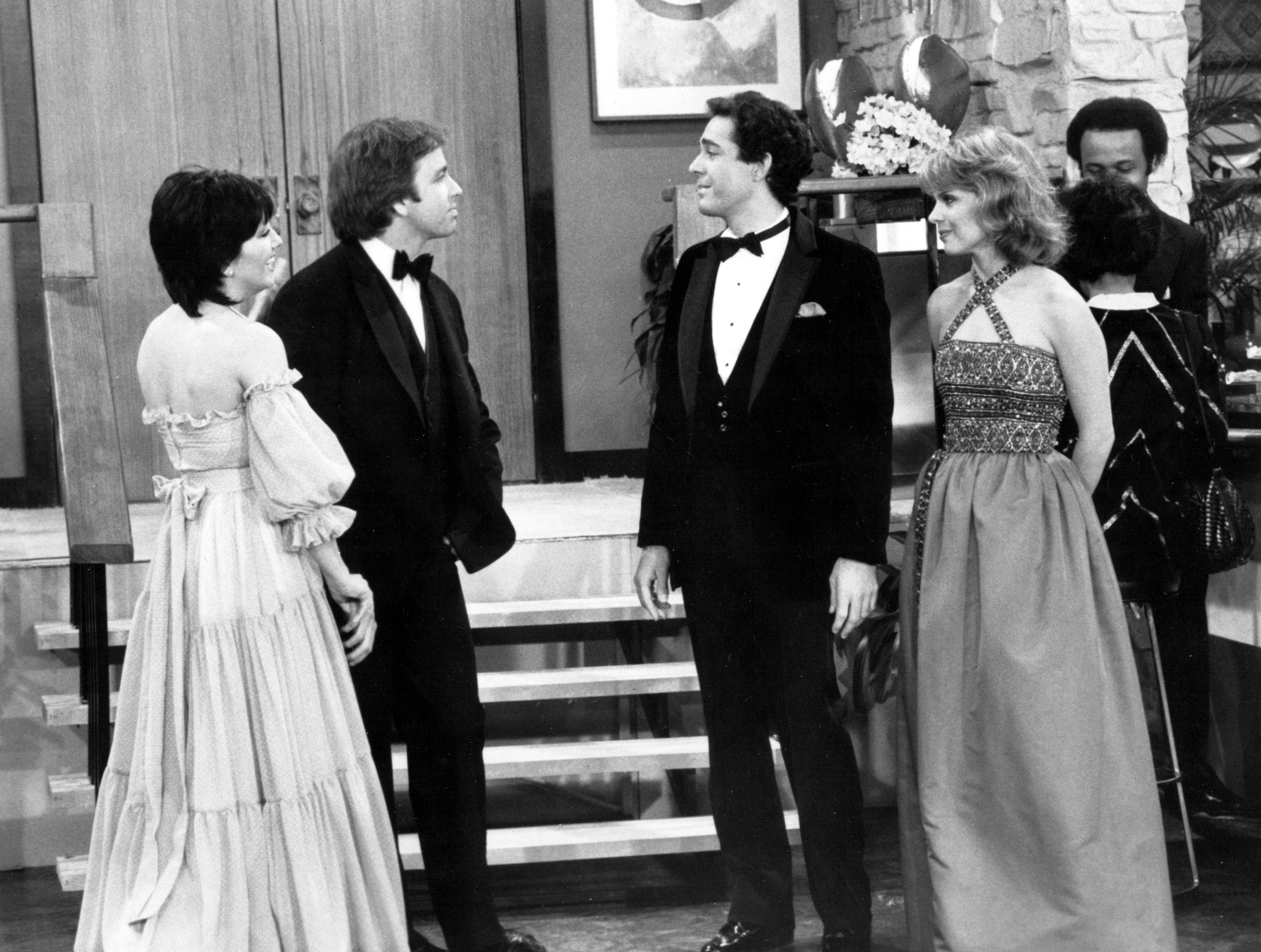 shot from the 1982 "Up in the Air" episode of "Three's Company." Left-to-right in this black-and-white image are Joyce DeWitt, John Ritter, Barry Williams and Lauree Berger. They are at a formal gathering, with the women in evening gowns and the men in tuxedos.