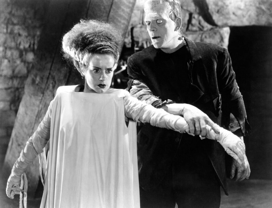 black-and-white image from the 1935 movie "The Bride of Frankenstein." On the left is Elsa Lanchester as the Bride, wearing a white shroud/gown, staring intently as she holds out her left arm, which is being held by Frankenstein's monster (Boris Karloff) on the right of the photo, who is looking at her with curiosity.