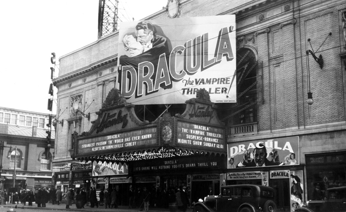 black-and-white image of the outside of the Stanley Theater in Jersey City, New Jersey, with its marquee advertising the showing of Bela Lugosi's film "Dracula." There are 1930s cars parked in front of the theater. A very large poster above the marquee has an illustration of Dracula about to bit a woman, with the hype copy: "Dracula, the Vampire Thriller!"