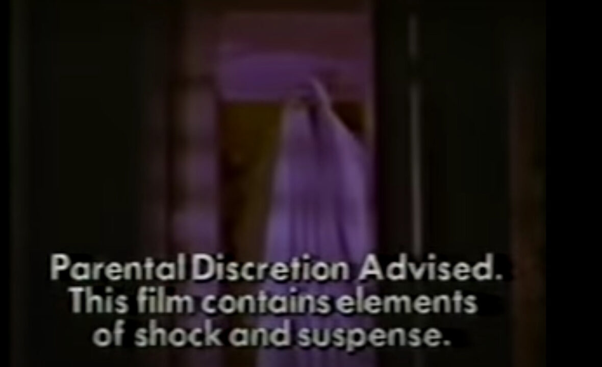 image from the intro to NBC's "Friday Night at the Movies" on Oct. 30, 1981, touting the premiere of the movie "Halloween." It depicts a scene from the movie in which the killer is disguised as a ghost under a sheet and standing in a doorway. Superimposed over this scene, in white lettering, reads: "Parental Discretion Advised. This film contains elements of shock and suspense."