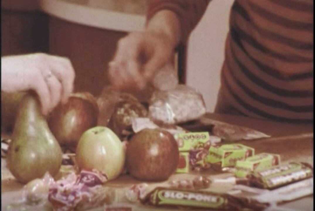 image from the 1977 short educational film "Halloween Safety." It depicts a girl and her mother at a kitchen table and going through the girl's treats she got while trick-or-treating, including several apples and some candy, to make sure they are safe to eat.