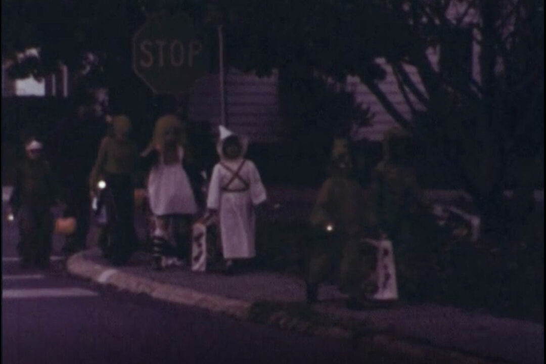 image from the 1977 short educational film "Halloween Safety." It shows a line of six children is costumes reaching the other side after crossing a street during a nighttime trick-or-treating session.