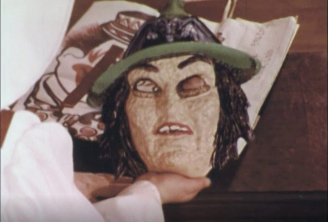 image from the 1977 short educational film "Halloween Safety." It shows the hands of a young girl holding the white-faced witch mask she will be wearing as part of her costume. Along with the face, the mask has a small witch's hat as part of it.