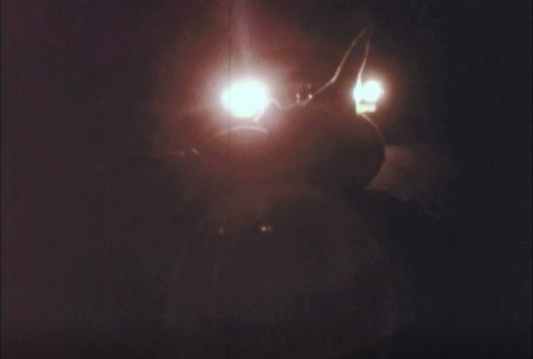 image from the 1977 short educational film "Halloween Safety." It is a grainy nighttime image, mostly dark except for the oncoming headlights of a car approaching the silhouette of a girl dressed in a witch's hat, who has fallen into the street because of her unsafe costume.