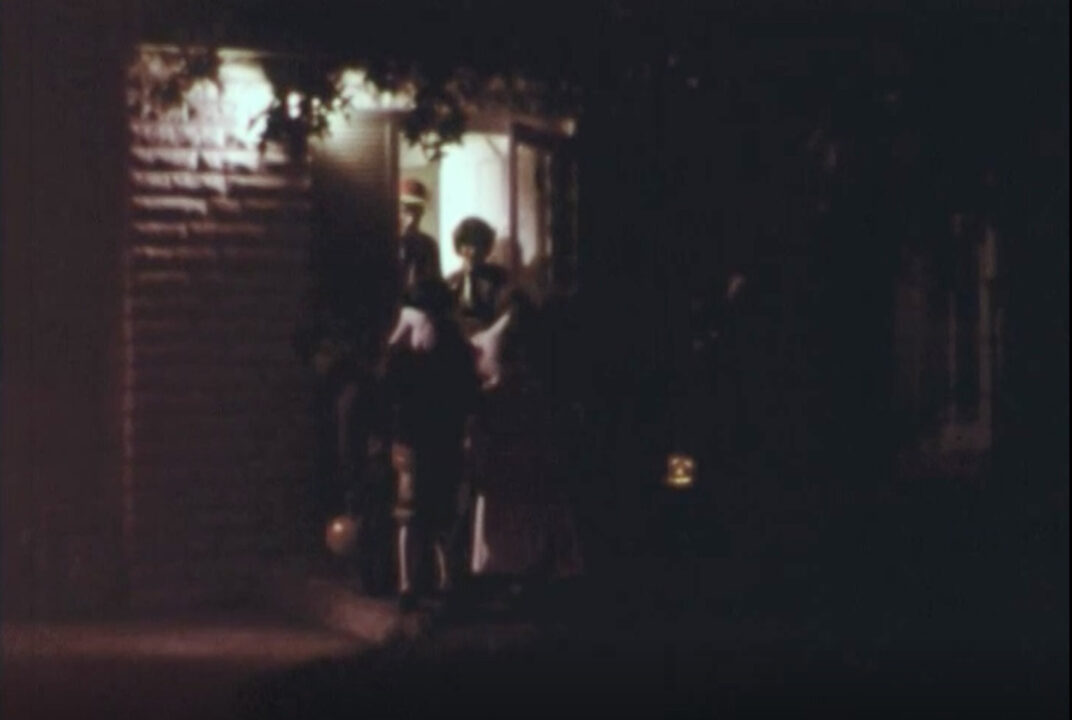 image from the 1977 short educational film "Halloween Safety." It is a nighttime view from a sidewalk looking at a house lit up for trick-or-treaters. A lit jack-o-lantern can be seen on the ground to the right of the porch, on which are standing several children in costumes receiving treats in their bags from the homeowners. 
