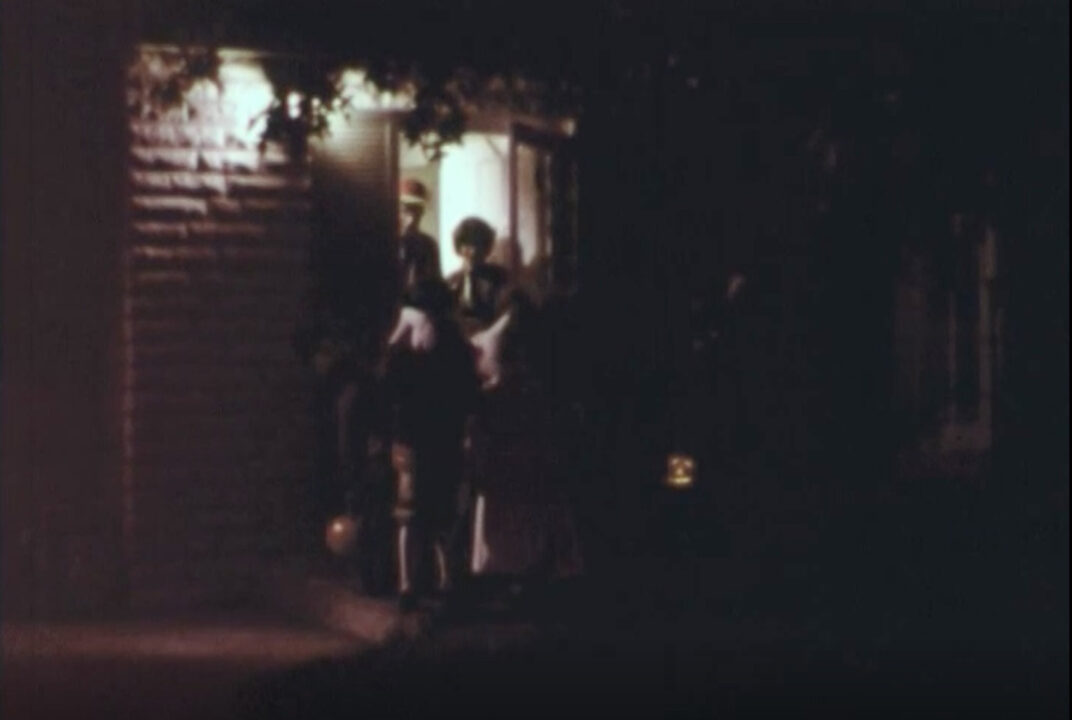 image from the 1977 short educational film "Halloween Safety." It is a nighttime view from a sidewalk looking at a house lit up for trick-or-treaters. A lit jack-o-lantern can be seen on the ground to the right of the porch, on which are standing several children in costumes receiving treats in their bags from the homeowners.