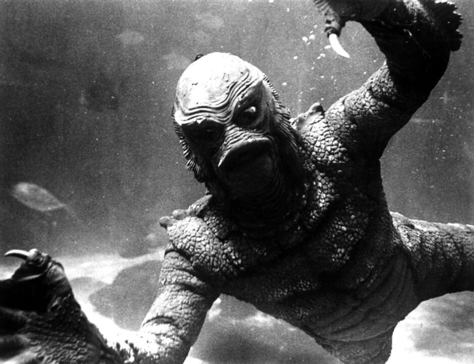 image from the 1955 movie "Revenge of the Creature." It is a closeup of the Gill-man (Ricou Browning) up against the glass of an aquarium, with his arms raised up.