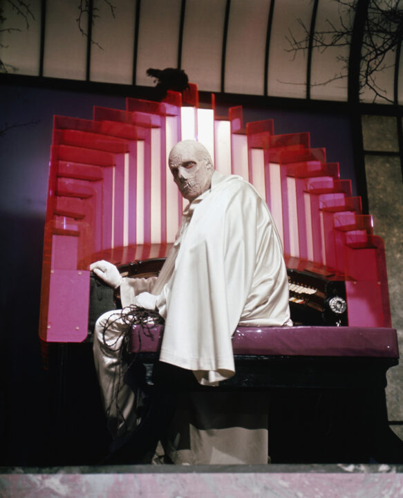 image from the 1971 film "The Abominable Dr. Phibes." It depicts Vincent Price as the title character, who is dressed all in white and wearing a mask that looks like a skull. He is seated at an organ, and looking away from the organ behind him toward the camera.
