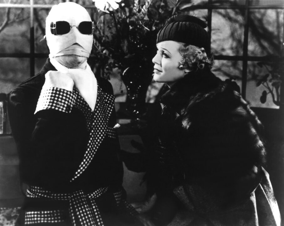 black and white image from the 1933 movie "The Invisible Man." Standing on the left of the photo is Claude Rains as the title character, wearing a dressing gown and with his head wrapped in bandages and wearing dark glasses. He is holding his right fist up in the air as if making a strong pronouncement. Directly to his left is costar Gloria Stuart, wearing a fashionable coat and hat, looking at him with concern.