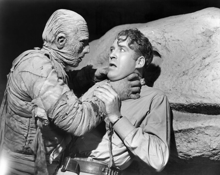 black and white image from the 1940 movie "The Mummy's Hand." on the left, the mummy Kharis is reaching up with his right hand and grasping the throat of an archaeologist, looking terrified on the right of the photo, with his back up against a stone wall.