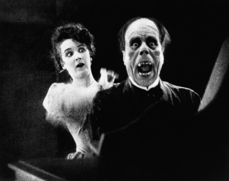 black and white image from the 1925 movie "The Phantom of the Opera." On the left of the photo is costar Mary Philbin, who has just unmasked the title Phantom, played by Lon Chaney. The Phantom is seated at an organ and has a look of surprise and anger on his deformed face after being unmasked.