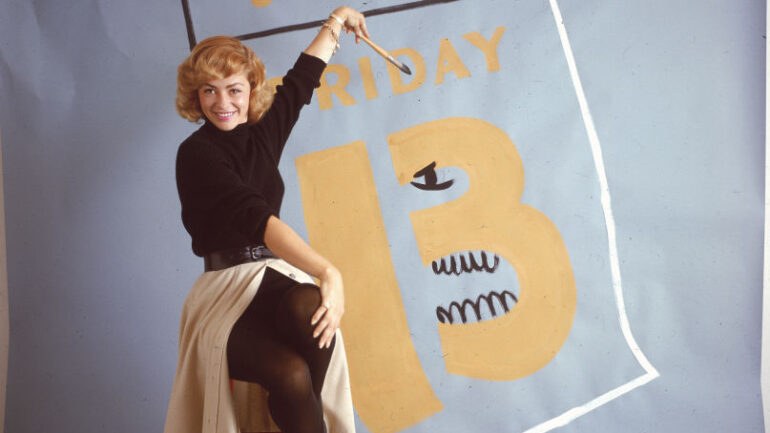 A woman smiles while sitting on a stool and painting a black eye and teeth on a large calendar page for 'Friday The 13,' circa 1965