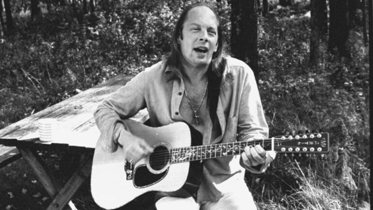 Author Rock Brynner sitting at picnic table, playing his guitar & singing, at home.