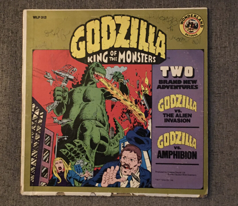 image of the album cover for the 1977 children's story record "Godzilla: King of the Monsters." It depicts an illustrated, comic-book-like image of the large green Godzilla trampling through a city as frightened people flee. He is holding a crushed bridge in his right hand (on left of illustration as Godzilla is approaching) and a destroyed jet plane in his left. He has his head turned toward his left and his breathing fire. Other jets are approaching to attack him. On the right side of this illustration, in a column with a purple background, reads: "Two Brand New Adventures; Godzilla vs. the Alien Invasion; Godzilla vs. Amphibion"