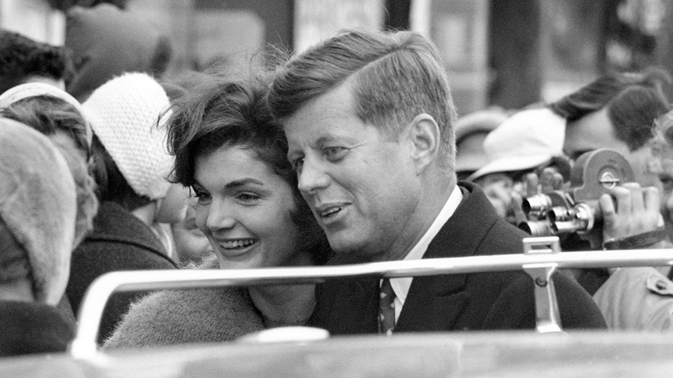 John F. Kennedy and Jacqueline Kennedy walk Main Street amongst crowd in Fort Atkinson, Wisconsin. White bar at bottom is roof lugagge rack rail atop station wagon. 1960