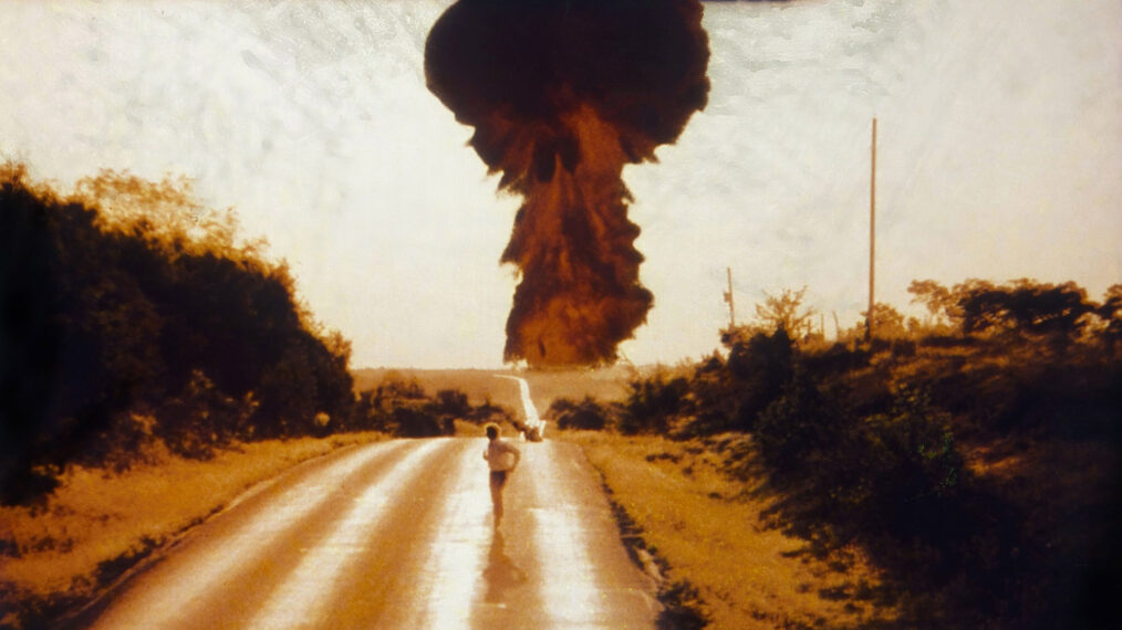 image from the 1983 TV movie 'The Day After.' It depicts a boy running toward the camera along a deserted country highway. In the background, looming over the horizon, is a mushroom cloud following a nuclear strike.