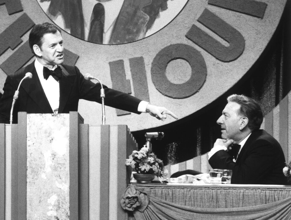 black and white image from a 1978 "Dean Martin Celebrity Roast" special. On the left of the photo, standing at a lectern and wearing a tuxedo, is actor Tony Randall. He is smiling and pointing down with his left hand at actor Jack Klugman, the roastee, who is seated on the right of the photo and also wearing a tux, and laughing.