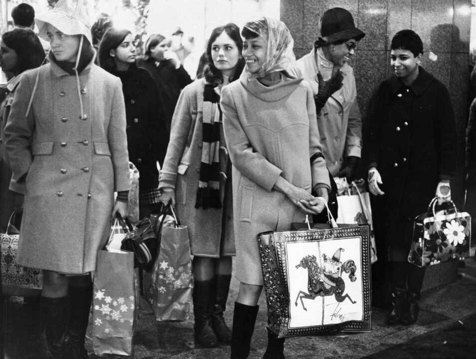black and white 1968 photo showing Christmas shoppers in Boston doing last minute shopping on Dec. 23. At the center of the photo are three women smiling and carrying Christmas-themed shopping bags.