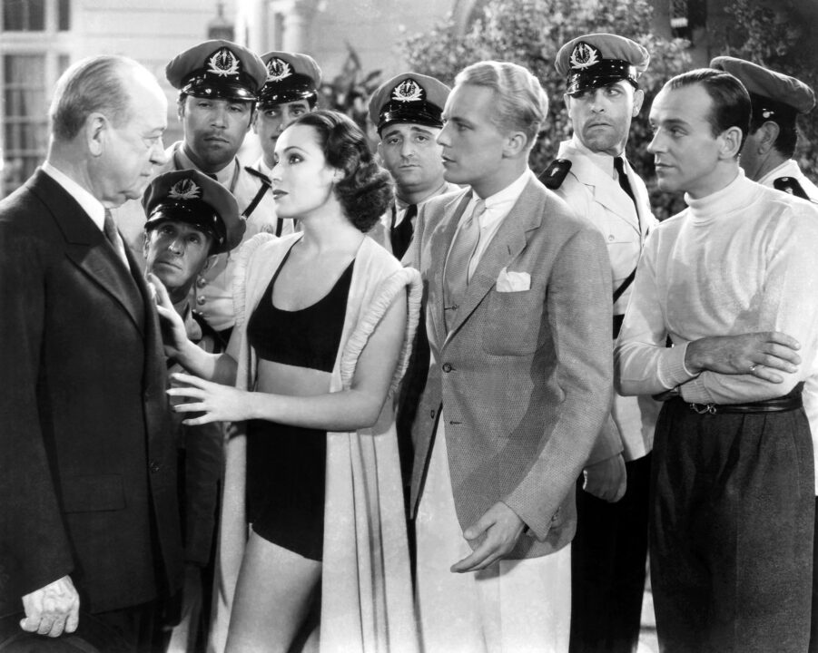 black and white image from the 1933 movie "Flying Down to Rio." Left to right are stars Walter Walker, Dolores del Rio, Gene Raymond and Fred Astaire. del Rio is wearing a black two-piece women's bathing suit, the first major actress to wear one onscreen (a white wrap is draped over her shoulders).