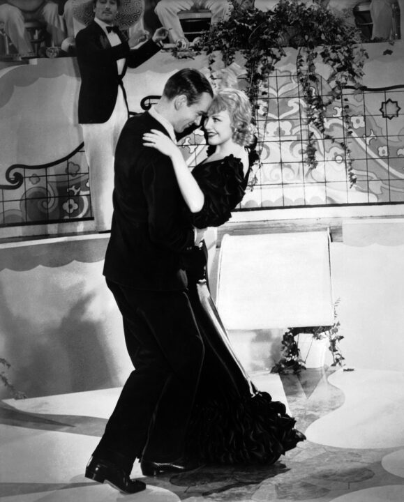 black and white image from the 1933 movie "Flying Down to Rio." It is the scene where Fred Astaire and Ginger Rogers dance together onscreen for the first time, accompanied by the song "Carioca." In this vertical image, Astaire is on the left, wearing a dark tuxedo. Rogers is wearing a dark-colored ballgown, and is dancing very closely to Astaire; so close that their foreheads are touching. They are both smiling and looking into each other's eyes, with Astaire crouching down a little mid-dance and because he is about a head taller. In the background, a bandleader is conducting musicians.