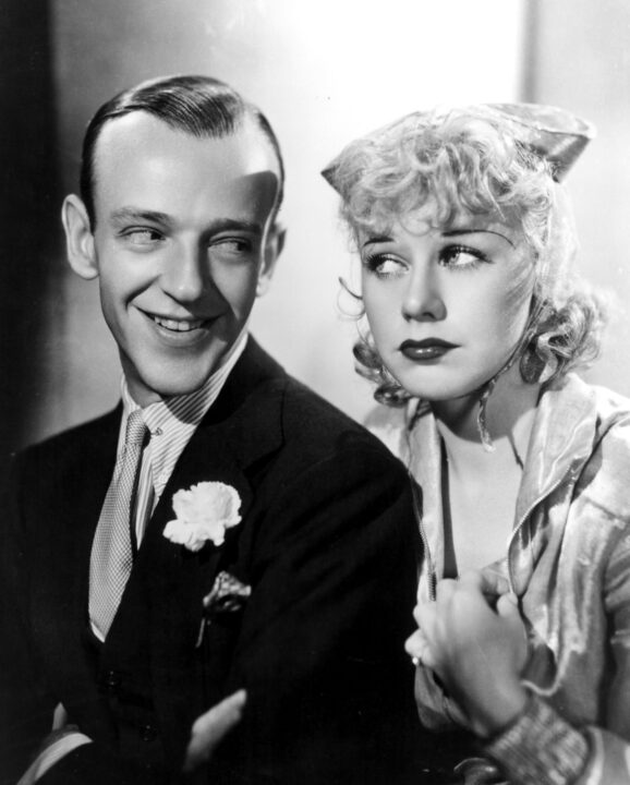 black and white photo from the 1933 movie "Flying Down to Rio." It is a medium shot of stars Fred Astaire (on left) and Ginger Rogers in character. Astaire is smiling and wearing a dark suit with a white carnation in the pocket, and a lighter colored dress shirt and tie. Rogers is wearing a lighter colored dress and a head covering tied around her neck.