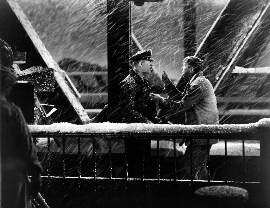 black-and-white image from the 1946 movie "It's a Wonderful Life." It is a nighttime scene on a bridge in a heavy snowfall. George Bailey (James Stewart) is on the right of the photo, excitedly talking to Bert the cop (Ward Bond) on the left.