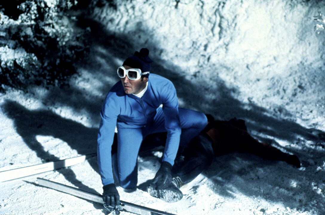 image from the 1969 James Bond movie "On Her Majesty's Secret Service." It is a nighttime shot, set in a snowy, alpine area, with George Lazenby as Bond wearing a ski outfit, skis and goggles, crouching down and hiding from bad guys pursuing him down the mountain.
