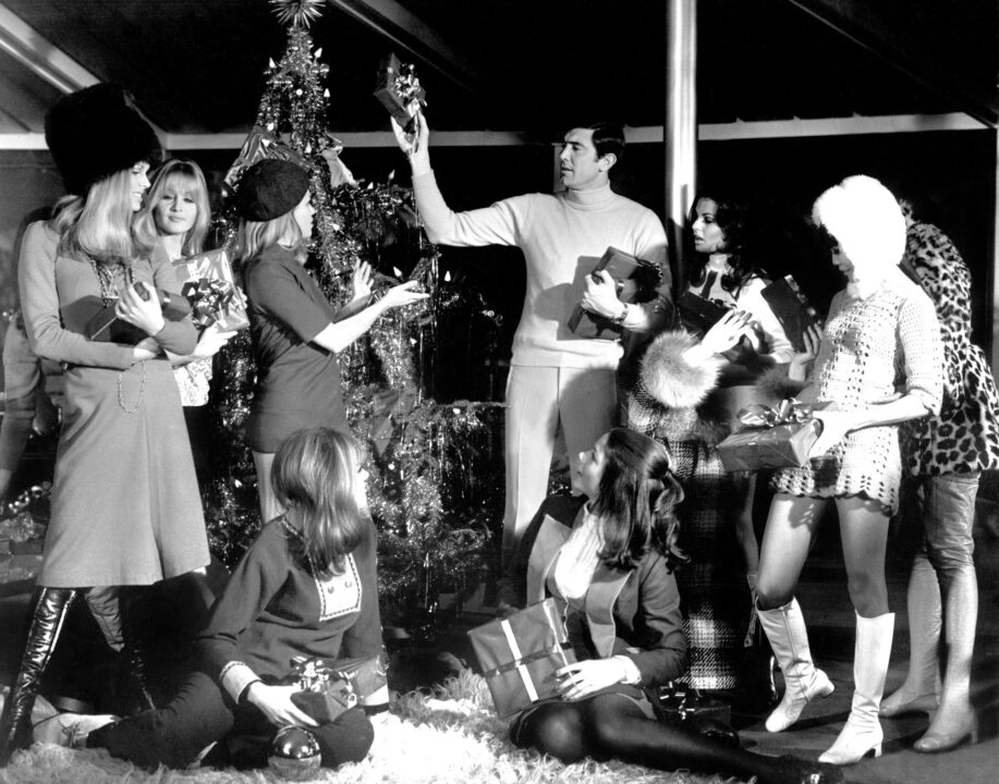 black-and-white image from the 1969 James Bond movie "On Her Majesty's Secret Service." Bond (George Lazenby) is in the center of the scene, wearing a '60s turtleneck sweater and tight slacks. He is holding a wrapped Christmas gift and a hardcover book in his left hand and is turned toward a decorated Christmas tree, upon which he is placing another wrapped gift with his left hand. He is surrounded by a group of young women of different ethnicities/nationalities who are dressed in chic '60s winter attire, including fur hats and boots.