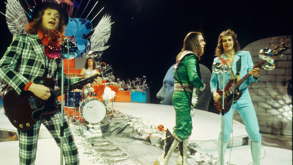 image of British rock group Slade performing on a Christmas TV show in the Netherlands in December 1973. Left to right are members Noddy Holder, Don Powell, Dave Hill and Jim Lea, dressed in glam rock attire of the era, including flashy bright green suits and tall boots.