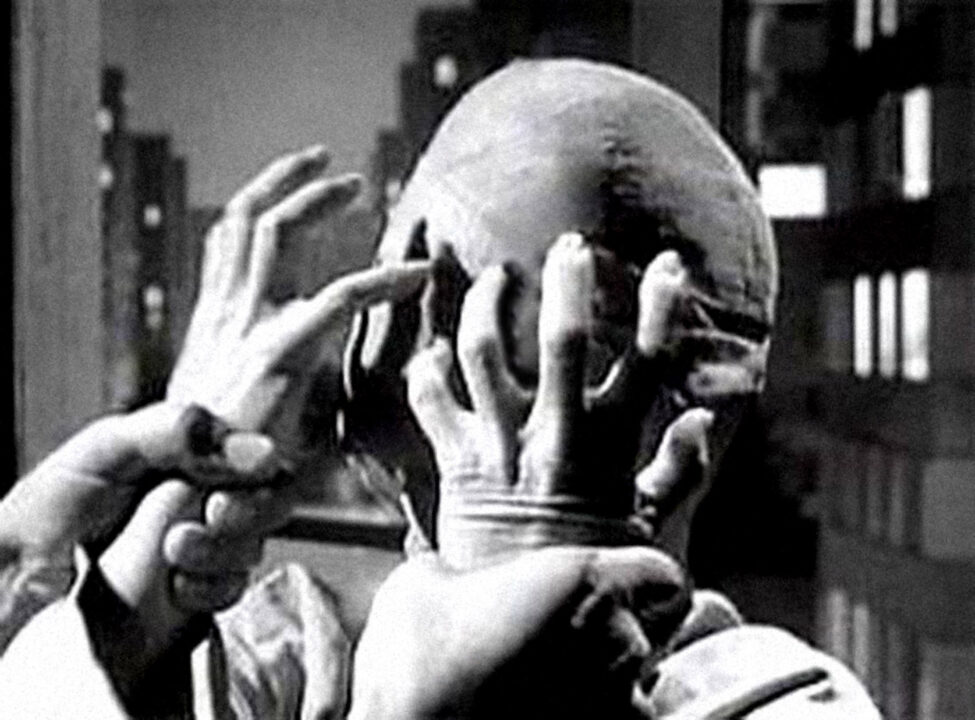 black and white image from the "Eye of the Beholder" episode of "The Twilight Zone." It is a closeup of a woman whose entire head and face is wrapped in bandages. She is reaching up as if desperately wanting to tear the bandages off, while a pair of another person's hands are grasping her wrists and holding them back.